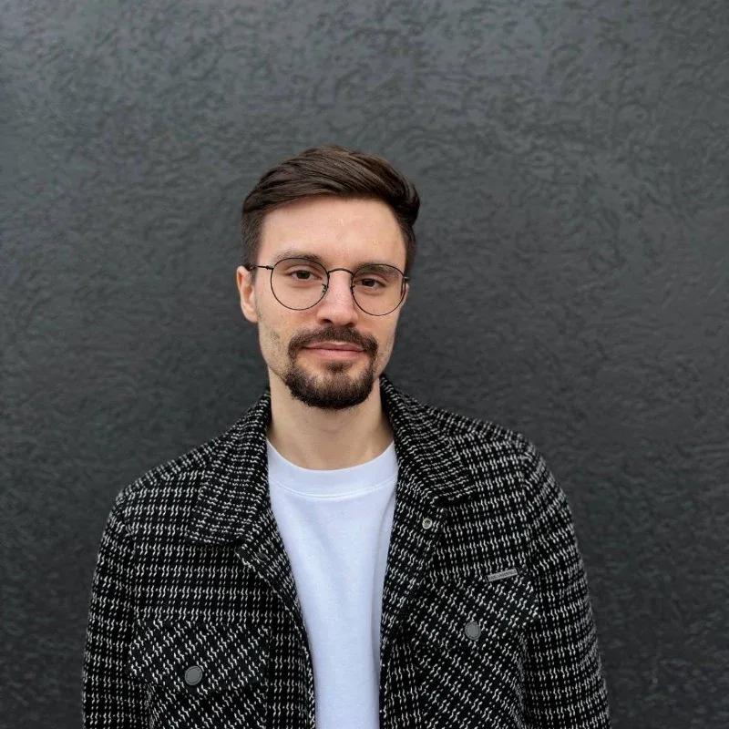 Oleg Chystieiev, in round glasses and a checked jacket over a white T-shirt, against a dark textured wall.